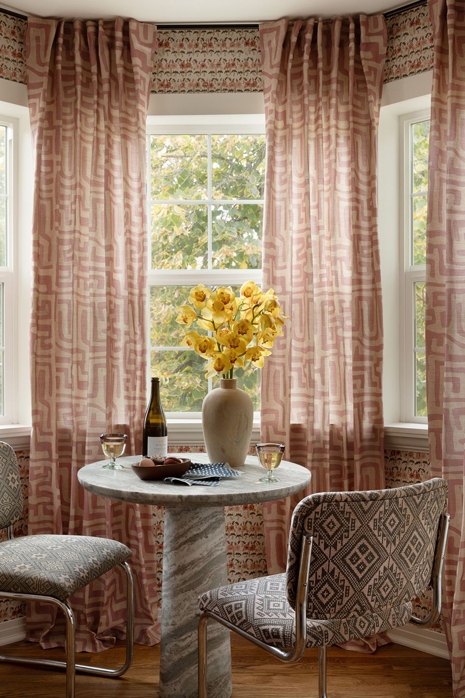 Bay window sitting area with mauve plaid curtains, patterned roman shade, and geometric upholstered chairs in warm, sophisticated tones.
