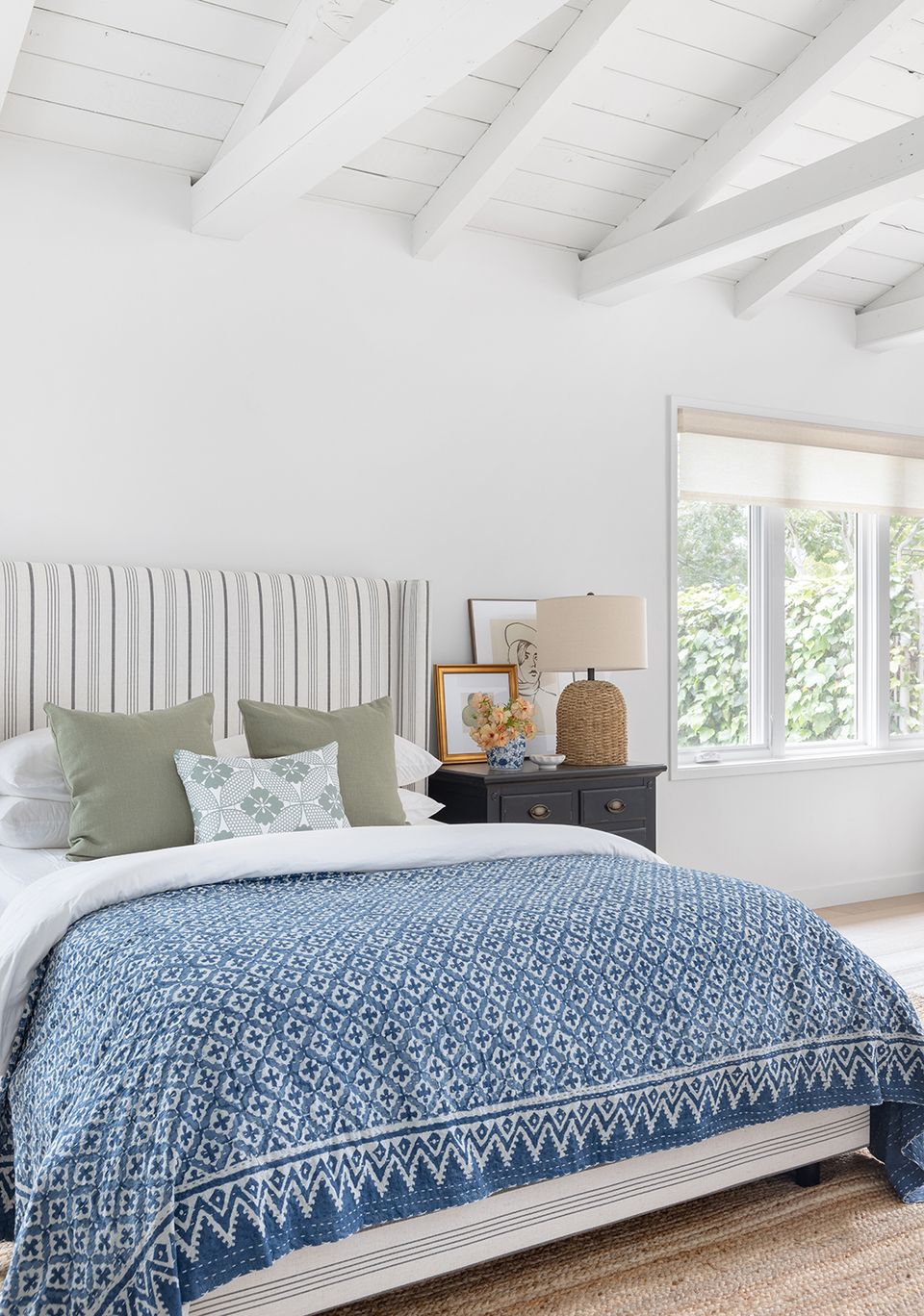 Bright coastal guest bedroom with ribbed headboard, blue patterned coverlet, and white beams