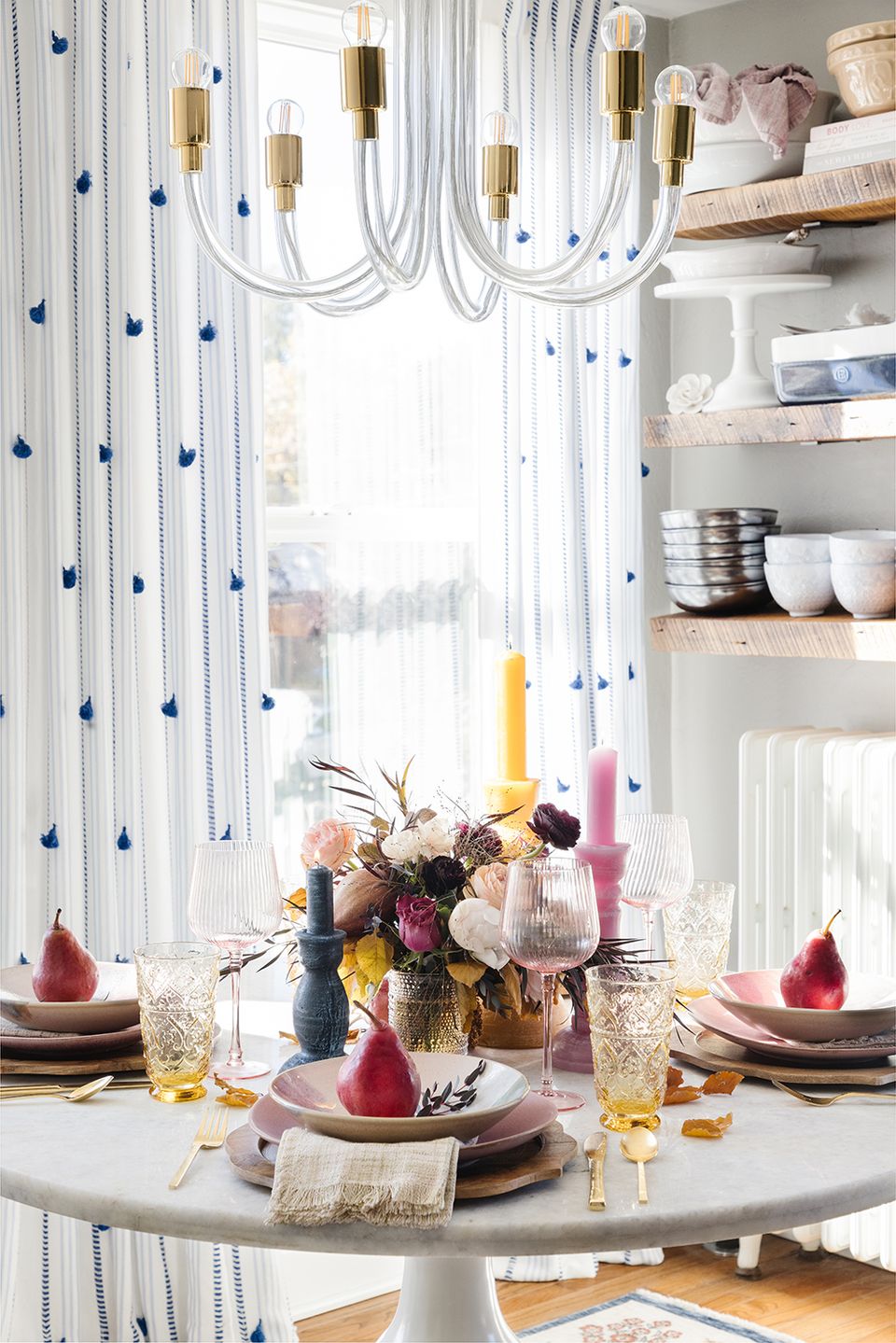 Whimsical dining setup with white curtains, blue teardrop pattern, brass chandelier, and colorful tablescape with pink pear accents