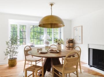 Organic modern dining room with brass pendant, natural wood table, woven chairs, and abundant greenery.