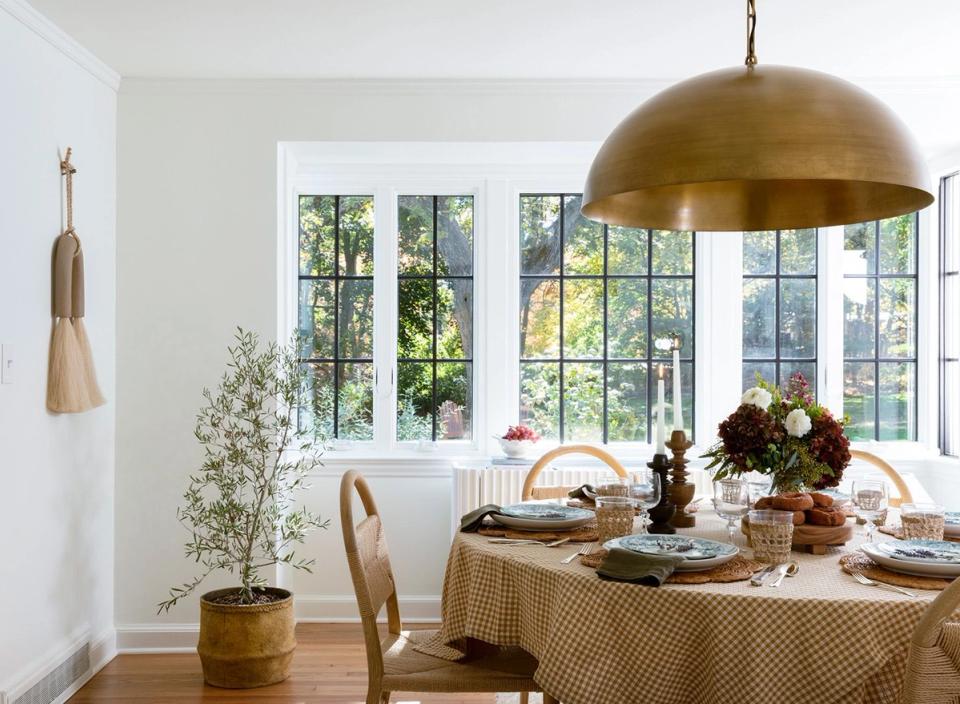 Warm, transitional dining room with brass pendant, neutral patterned tablecloth, and abundant natural light from gridded windows