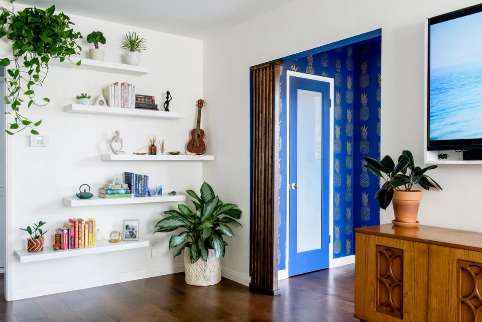 Bright living room with white floating shelves, bold cobalt blue door, warm wood furniture, lush greenery, and eclectic coastal-modern style.