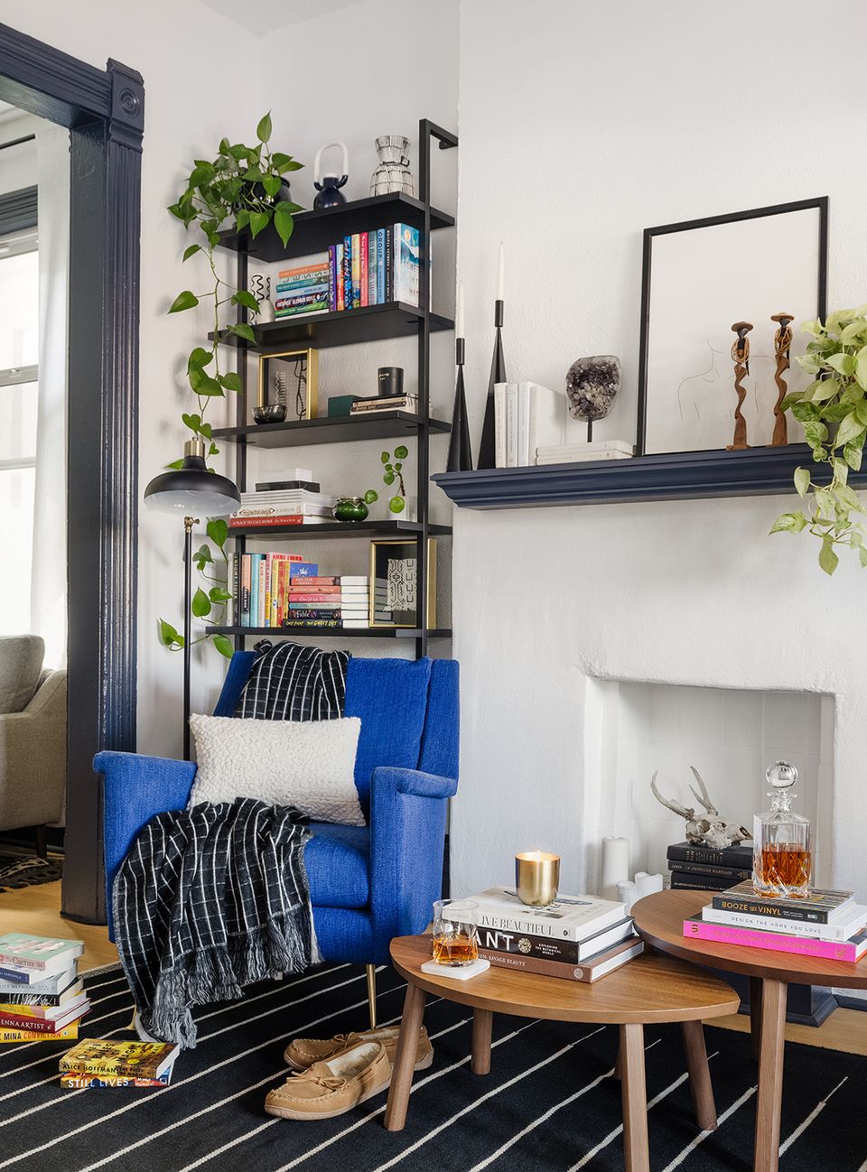 Eclectic bohemian corner with cobalt blue armchair, black metal shelving styled with books and trailing plants, mid-century wood table, and dark striped rug