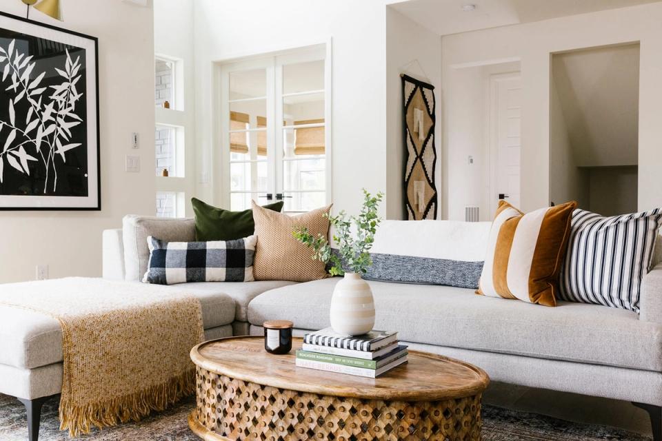 Contemporary neutral living room featuring gray sectional with mixed-pattern pillows in mustard, blue, and cream, and round wood coffee table