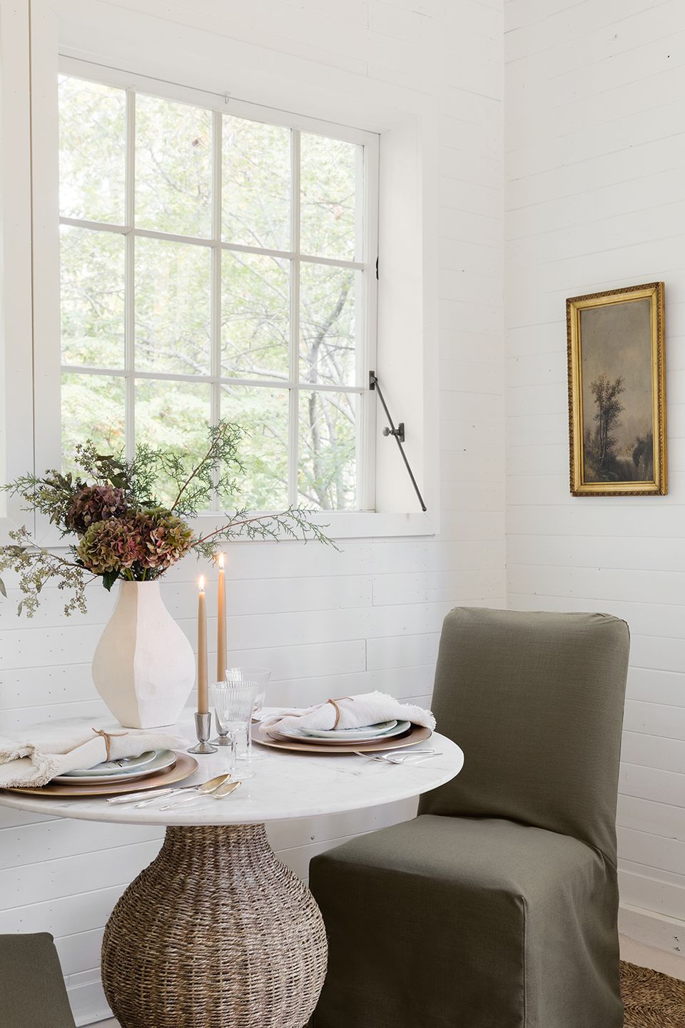 Modern dining nook with white round table, olive green chair, gold-framed painting, and dried flower arrangement by window