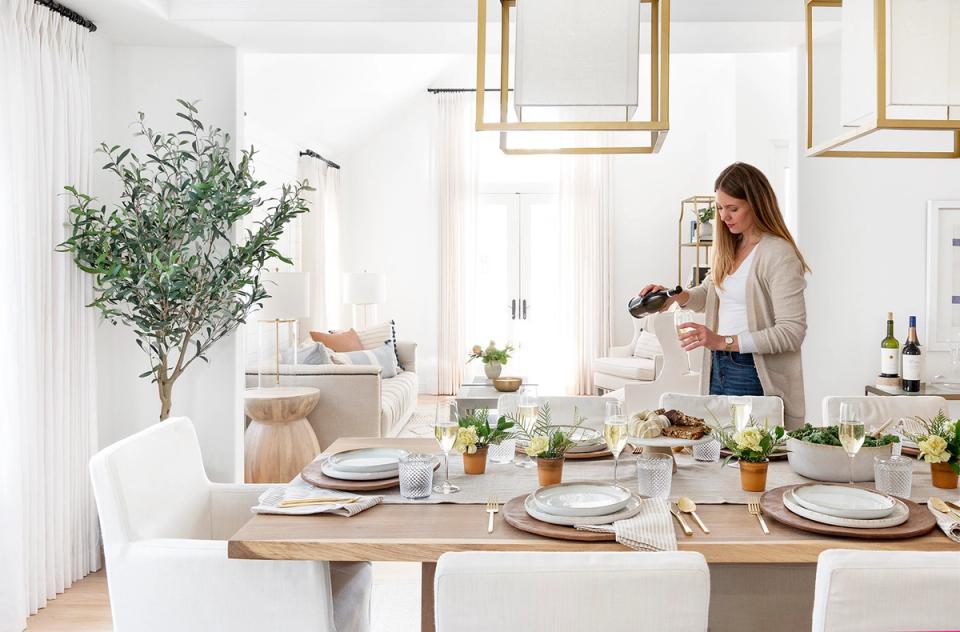 Bright minimalist dining room with natural wood table, white chairs, brass-framed mirrors, and greenery accents