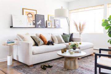 Contemporary neutral living room with floating shelves displaying art collection, cream linen sofa, and brass round side table