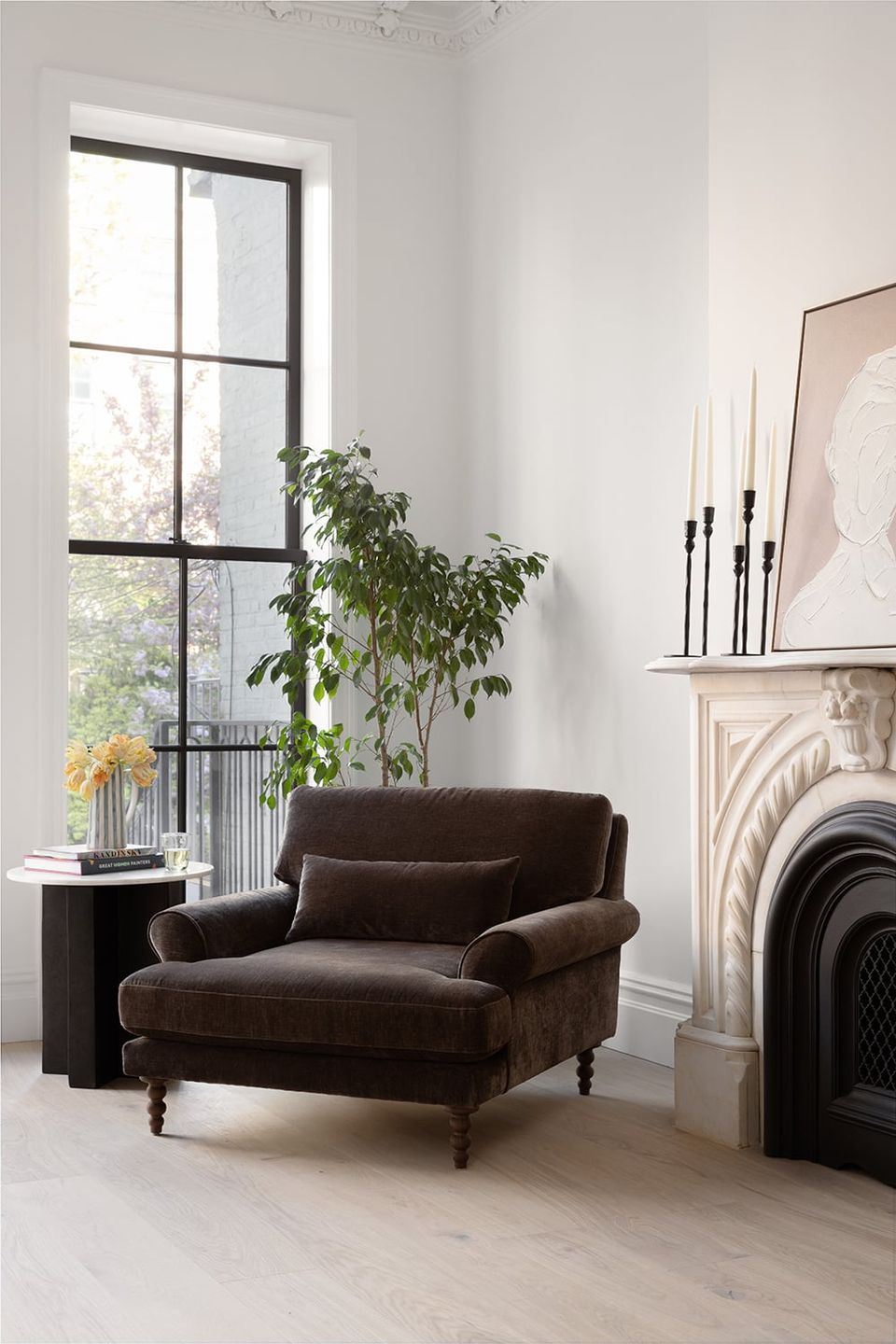 Minimalist modern living room with chocolate brown velvet chair, white walls, black-framed window, and fireplace mantel