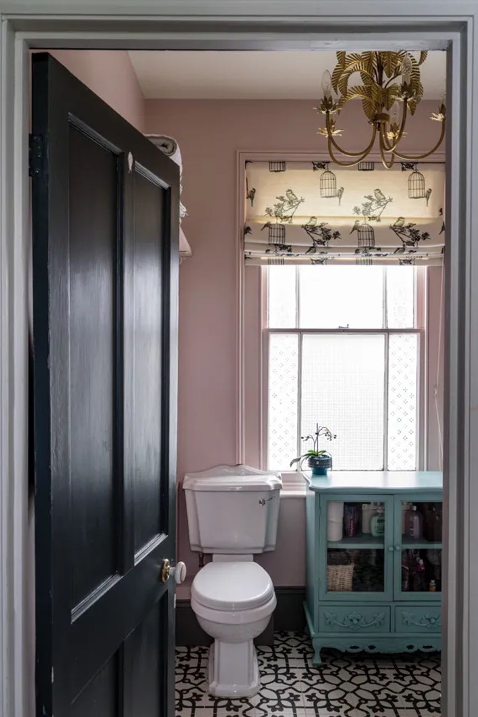 Soft pink bathroom with black door, turquoise vintage vanity, gold chandelier, and geometric tile floor creating elegant eclectic charm