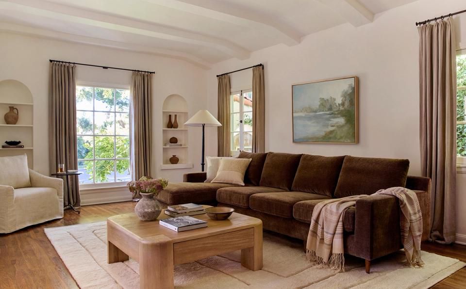 Inviting farmhouse-inspired living room with rich chocolate sectional, warm wood tones, soft taupe curtains, and natural textures creating a cozy, welcoming retreat.
