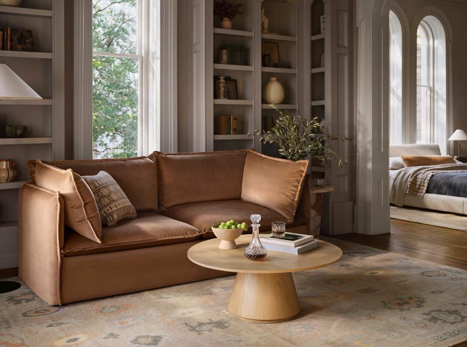 Warm transitional sitting area with cognac leather sofa, natural wood accents, soft neutral tones, and floor-to-ceiling shelving evoking a sophisticated, collected-over-time elegance.
