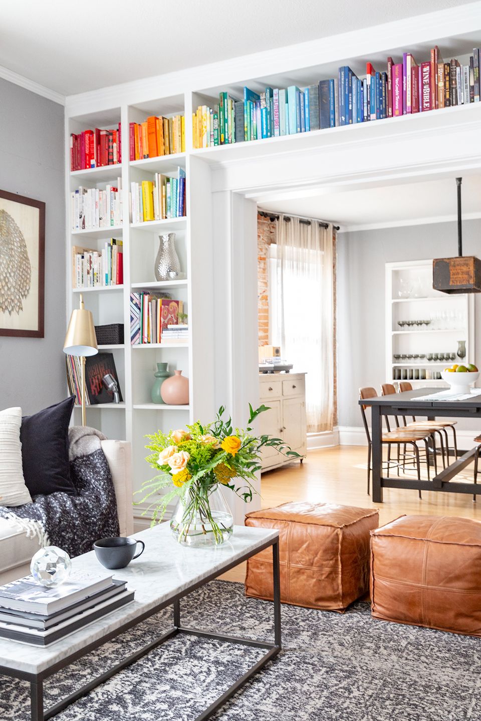 Contemporary living room featuring rainbow-organized built-in shelving as vibrant accent wall with cognac leather poufs.