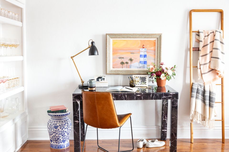 Minimalist workspace with mid-century modern touches: marble desk, cognac leather chair, brass lamp, and warm wood accents against white walls.
