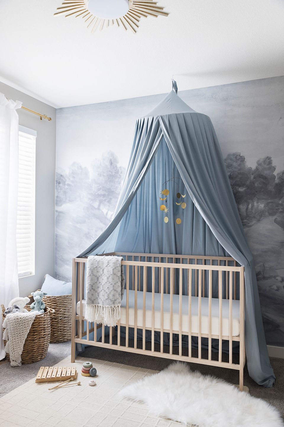 Soft nursery with blue canopy, nature mural backdrop, and gentle neutral tones with golden details