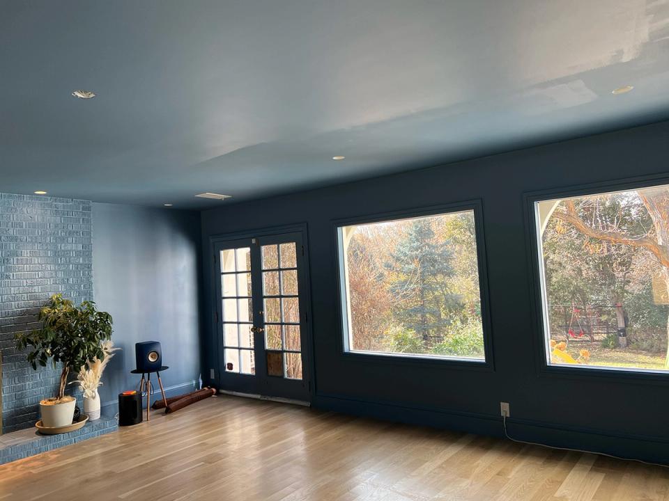 Minimalist room with deep teal walls, blonde wood floors, and expansive windows framing autumn views