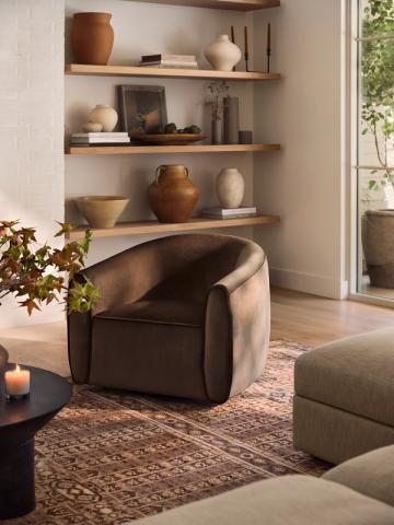 Earthy minimalist interior with warm wood shelving, curved charcoal armchair, pottery accents, and natural light creating serene ambiance.