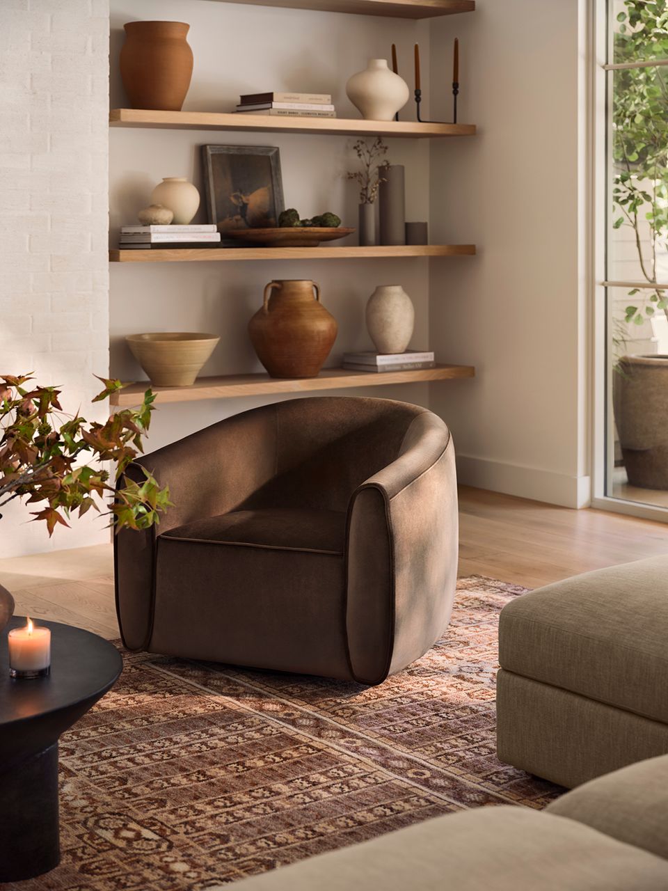 Earthy minimalist interior with warm wood shelving, curved charcoal armchair, pottery accents, and natural light creating serene ambiance.