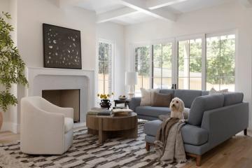 Serene modern living room with charcoal sectional, cream armchair, geometric rug, and abundant natural light from large windows.