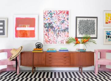 Eclectic gallery wall with vibrant floral prints and geometric art above mid-century wooden credenza, pink seating, bold black-and-white striped rug.