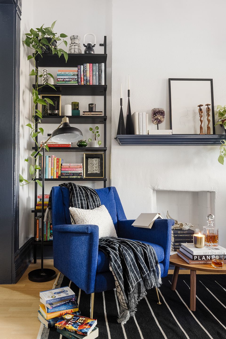 Modern reading nook featuring cobalt blue chair, black open shelving with layered styling, and green plants creating sophisticated, literary retreat