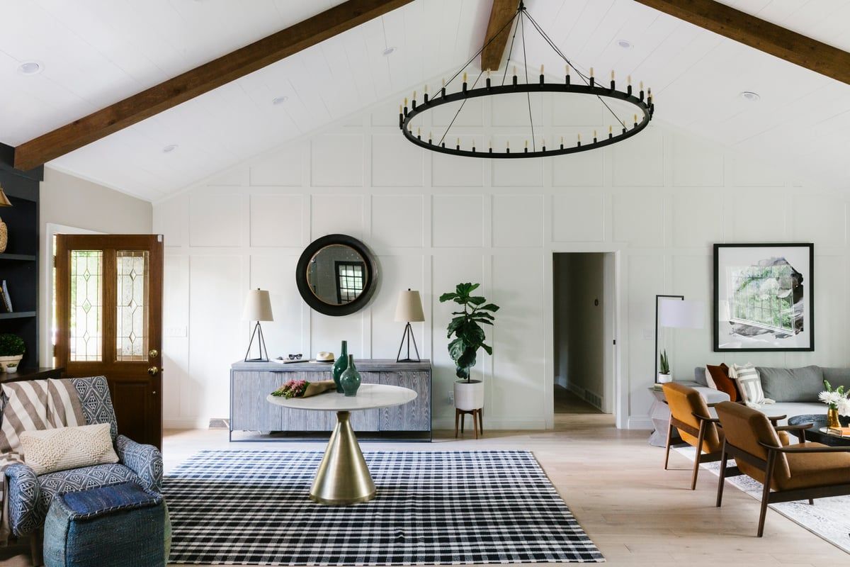 Bright open-plan living space with farmhouse aesthetic, white walls, wood beams, navy gingham rug, and mixed wood and upholstered furniture