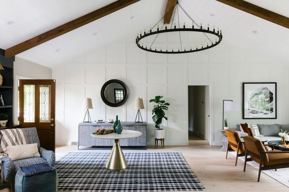 Bright open-plan living space with farmhouse aesthetic, white walls, wood beams, navy gingham rug, and mixed wood and upholstered furniture