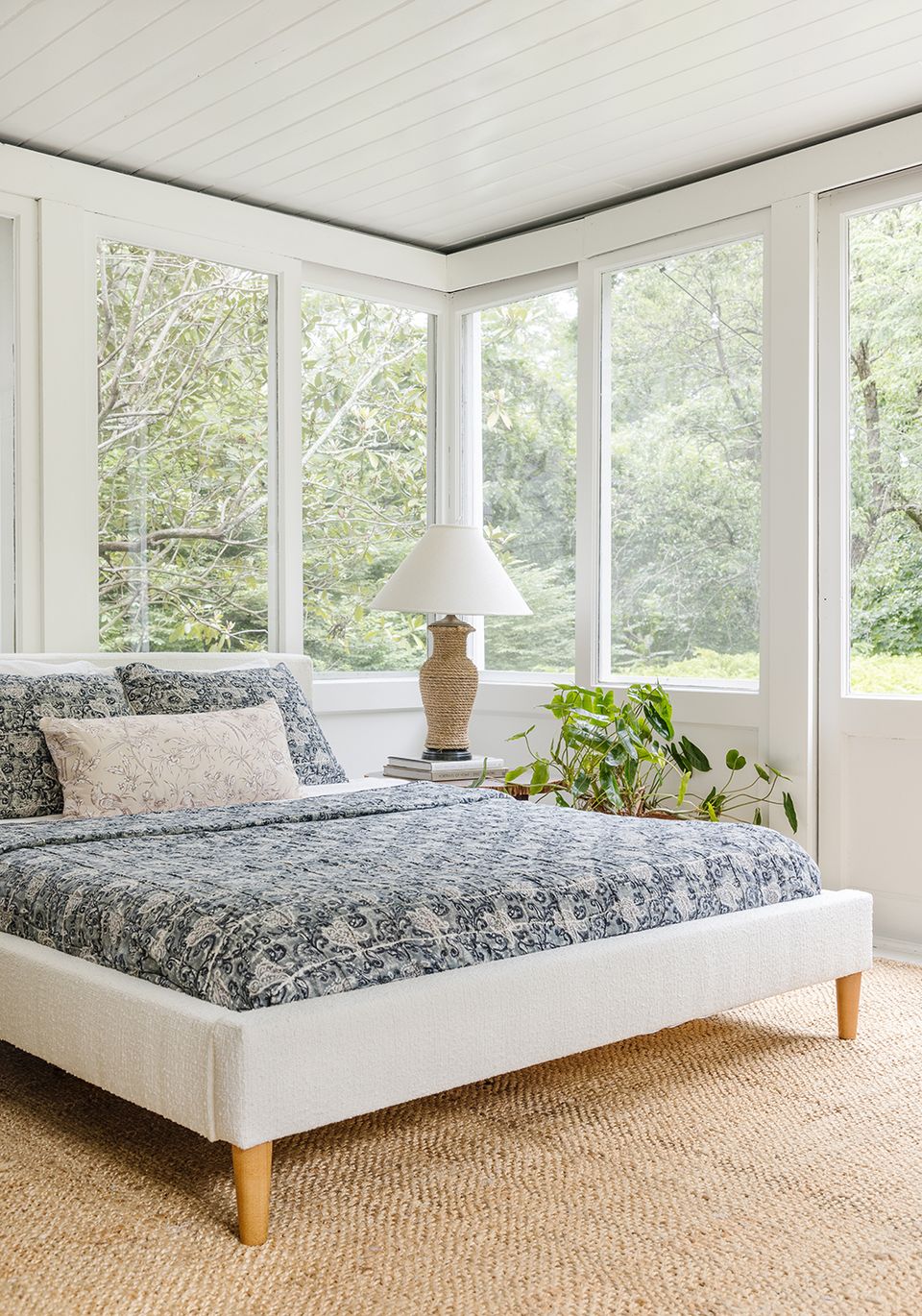 Serene sunroom bedroom with patterned bedding, natural wood legs, jute carpet, and lush green views from expansive windows