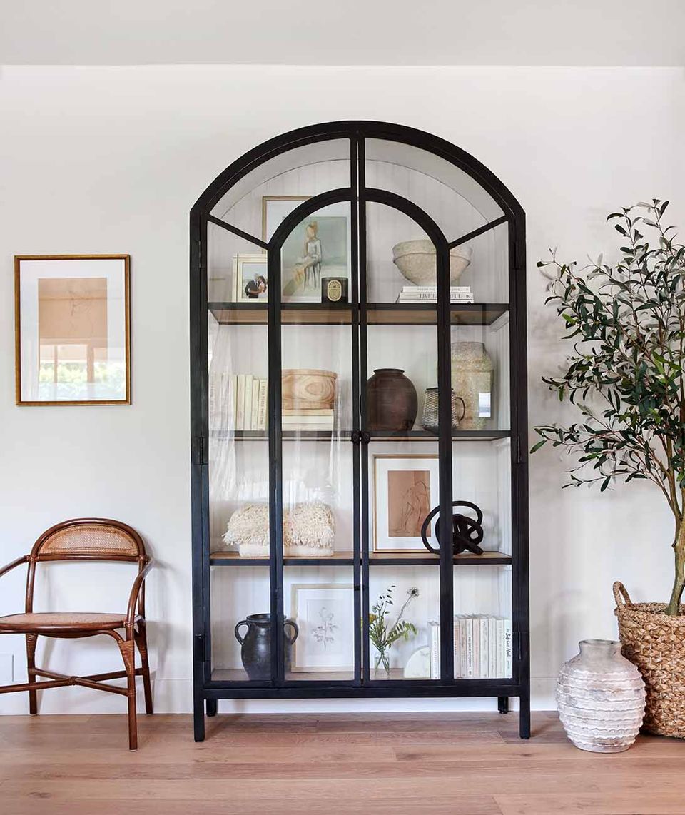 Contemporary traditional display cabinet: black arched vitrine with curated objects, wooden chair, potted plant, minimalist styling.