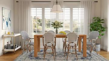 Airy coastal dining room with rattan chairs, wood table, shuttered windows, and lush plants