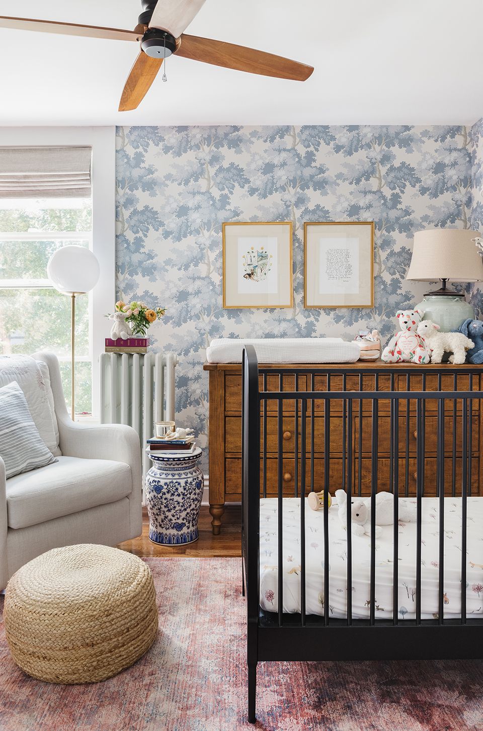 Modern nursery blending traditional style: blue toile wallpaper, wood dresser, black crib, neutral textures, and natural light.