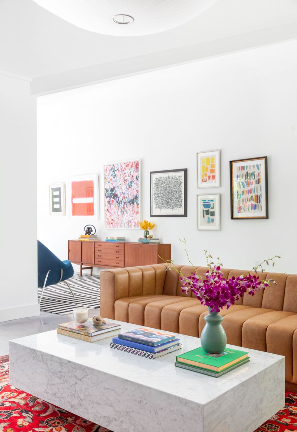 Mid-century modern living room: caramel sofa, colorful abstract prints, vintage wooden dresser, vibrant flowers, curated eclectic style.