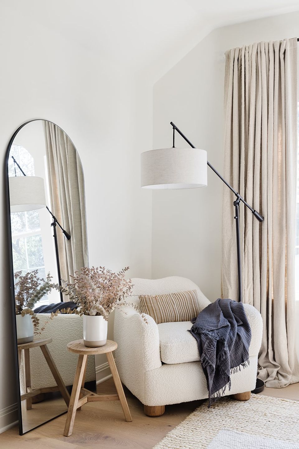 Minimalist bedroom corner with cream armchair, arched mirror, linen curtains, and pendant lamp creating serene, neutral aesthetic
