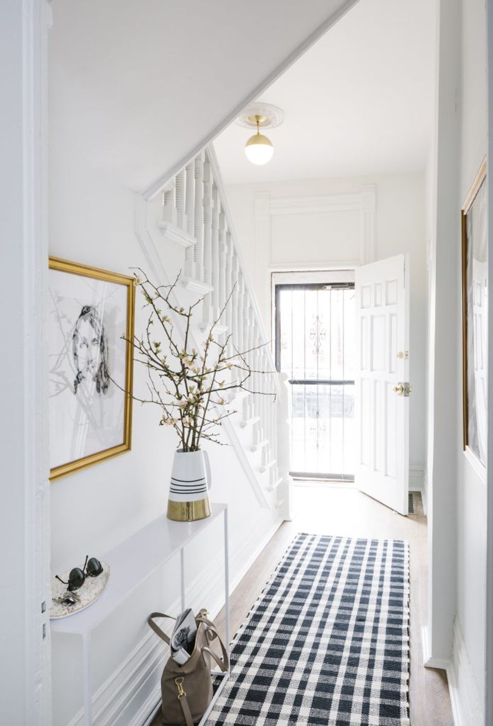 Bright minimal entry hallway with gold pendant light, framed botanical art, dried stems, and black-and-white checked floor runner