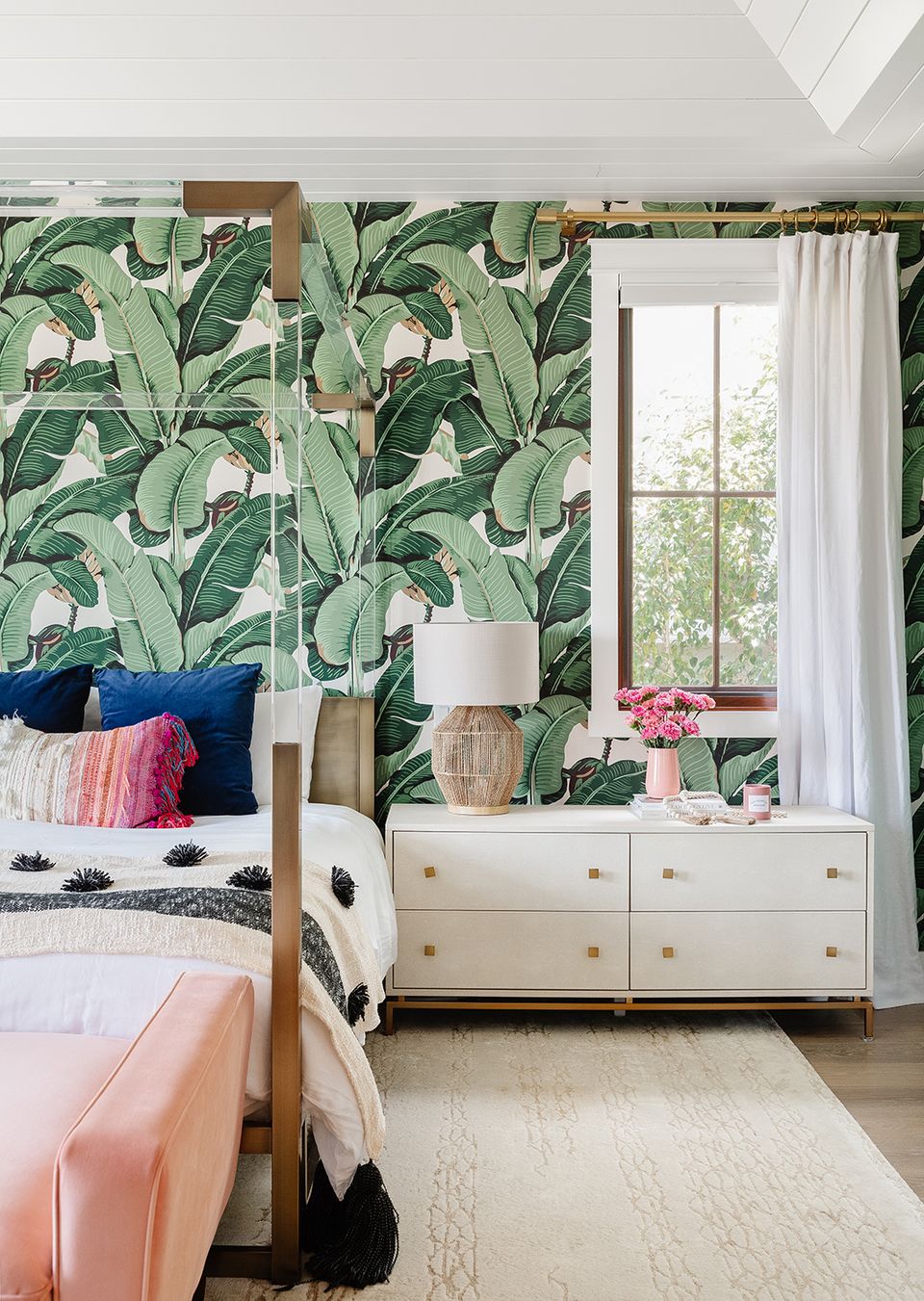 Tropical maximalist bedroom featuring bold banana leaf wallpaper, pink accents, and natural wood furnishings