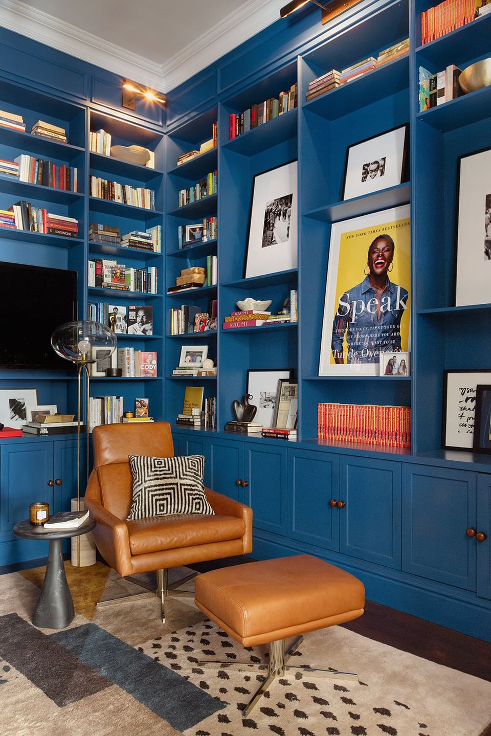 Bold eclectic office featuring deep blue built-in shelving, cognac leather seating, warm accent lighting, and vibrant book display