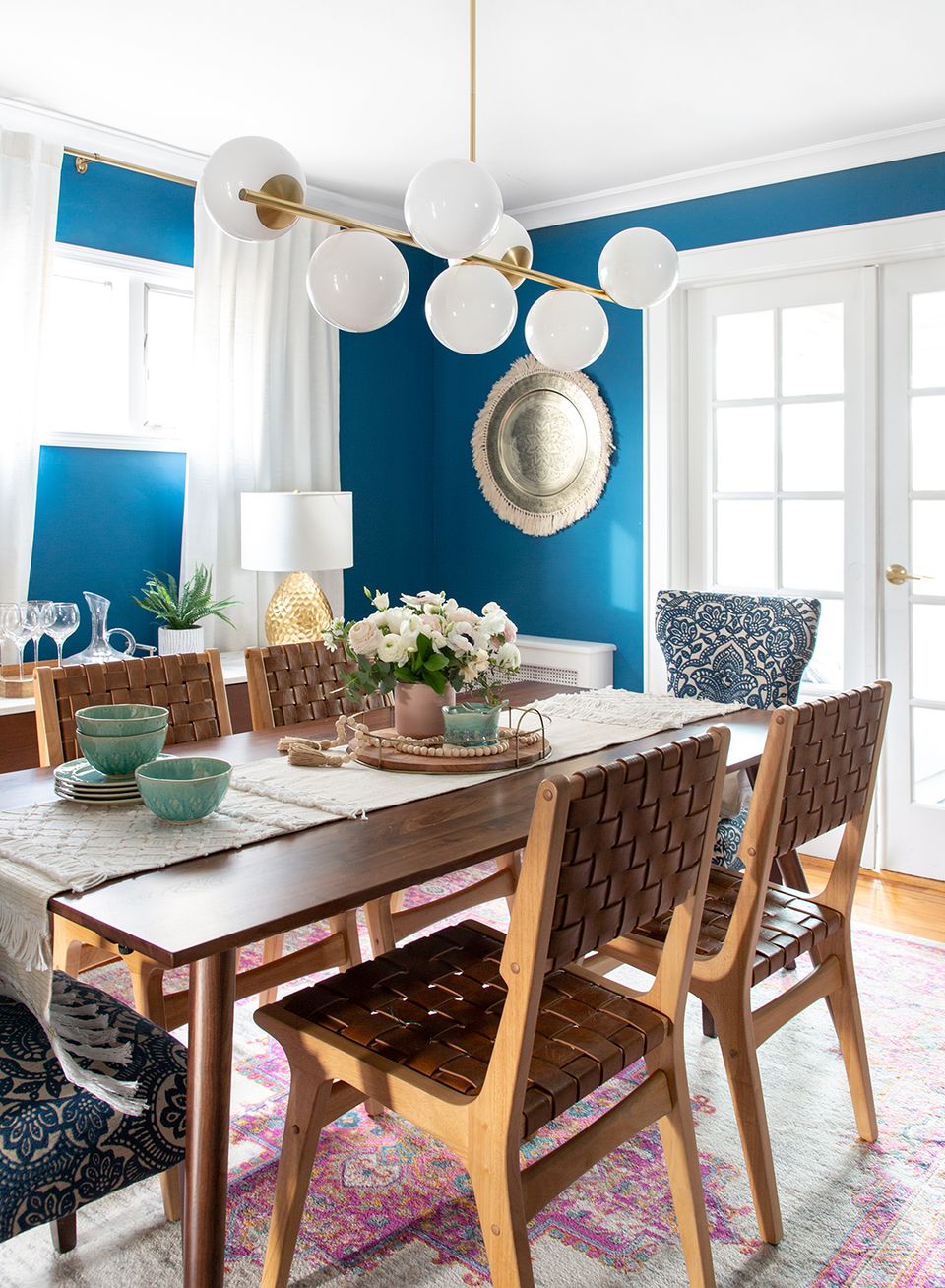Vibrant coastal dining space with bold teal accent wall, warm wood chairs, white globe pendant lighting, and layered textures in jewel tones.