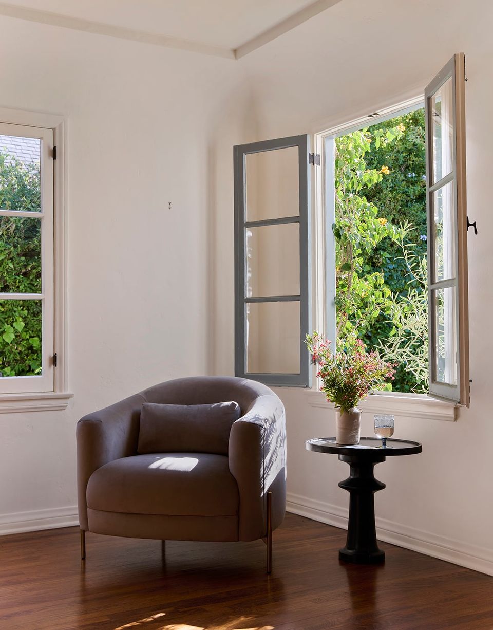Minimalist sunlit corner with chocolate brown rounded chair, black pedestal table, and expansive windows framing lush garden views