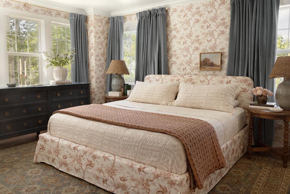 Traditional bedroom featuring floral wallpaper, gray drapery, and layered bedding in rose and cream tones