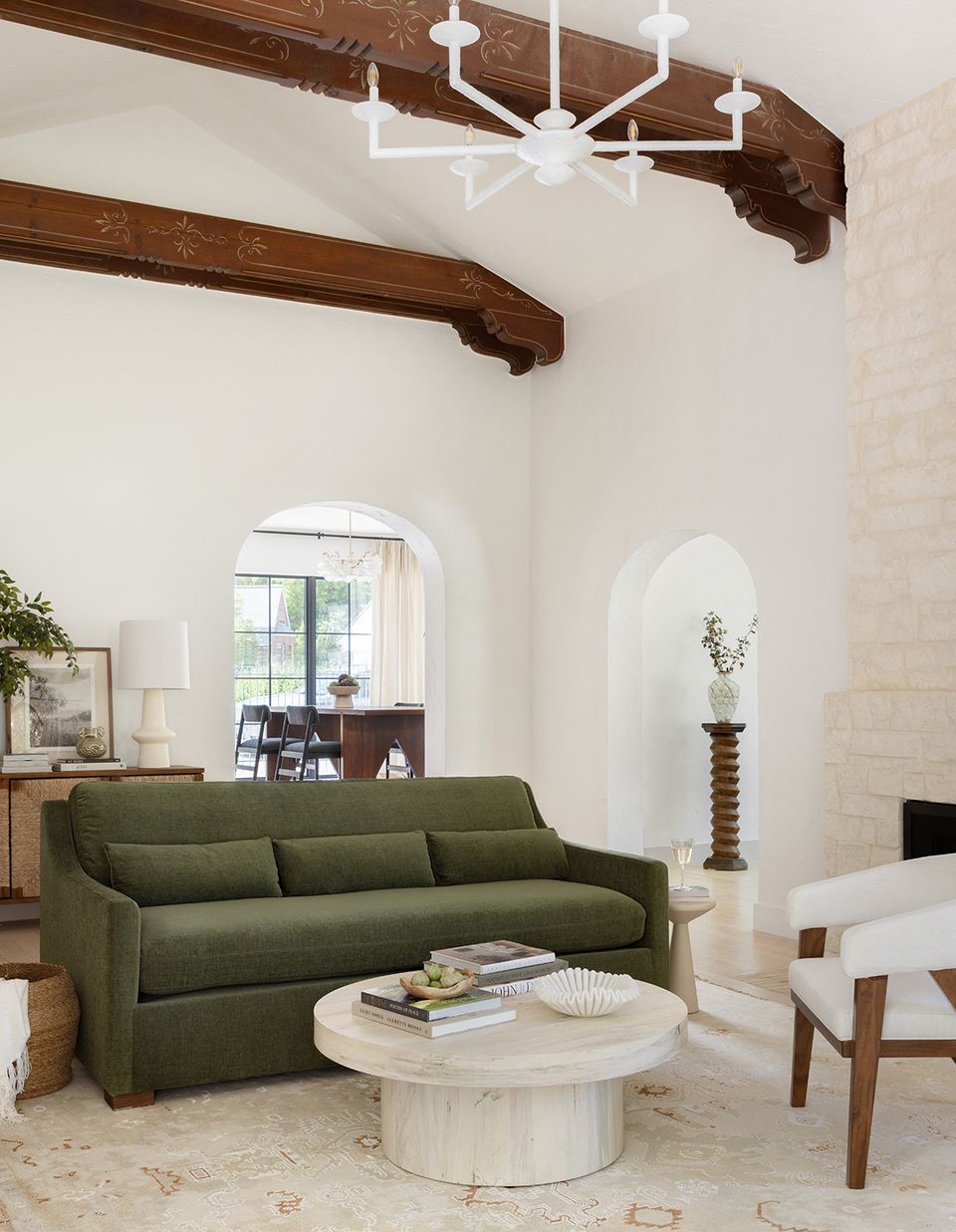 Modern rustic interior with curved sage green sofa, exposed wood beams, arched alcoves, and cream marble table for serene elegance