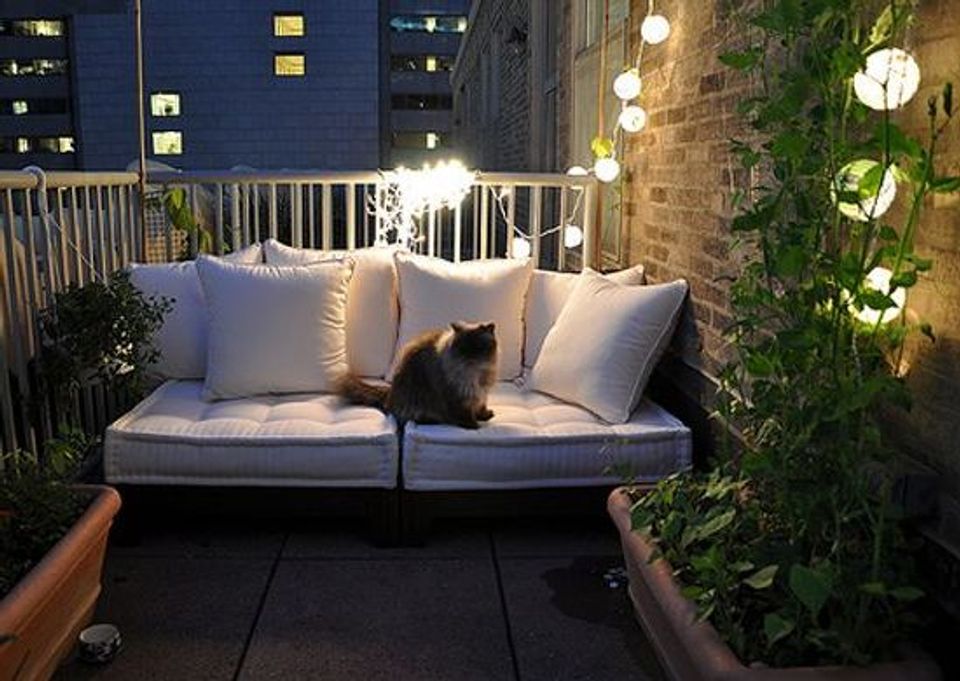 Modern minimalist balcony featuring a gray outdoor loveseat, white pillows, warm string lighting, and climbing ivy vines against dark urban backdrop.
