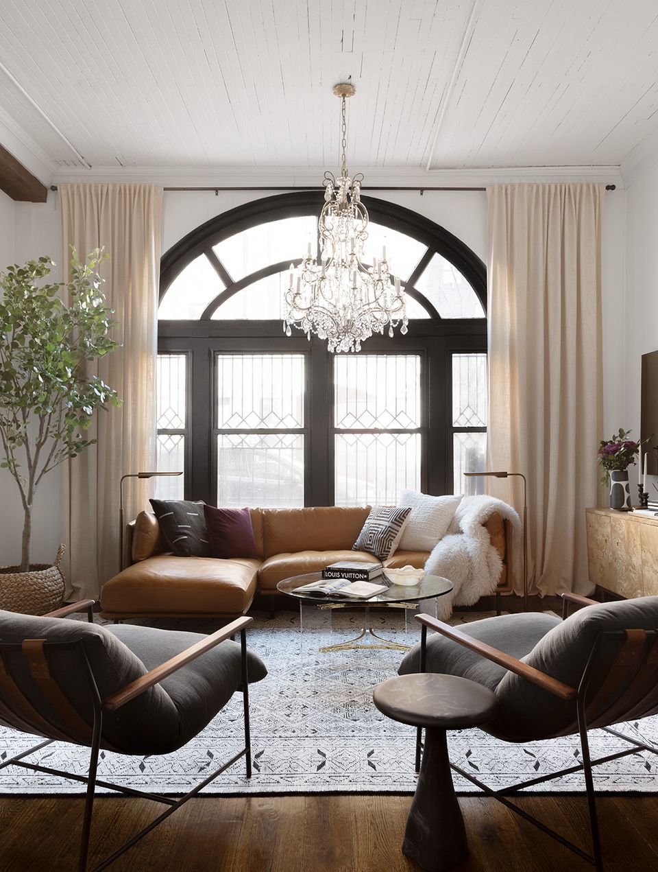 Contemporary seating area with caramel leather sofa, paired black lounge chairs, crystal chandelier, and neutral palette with architectural arch window
