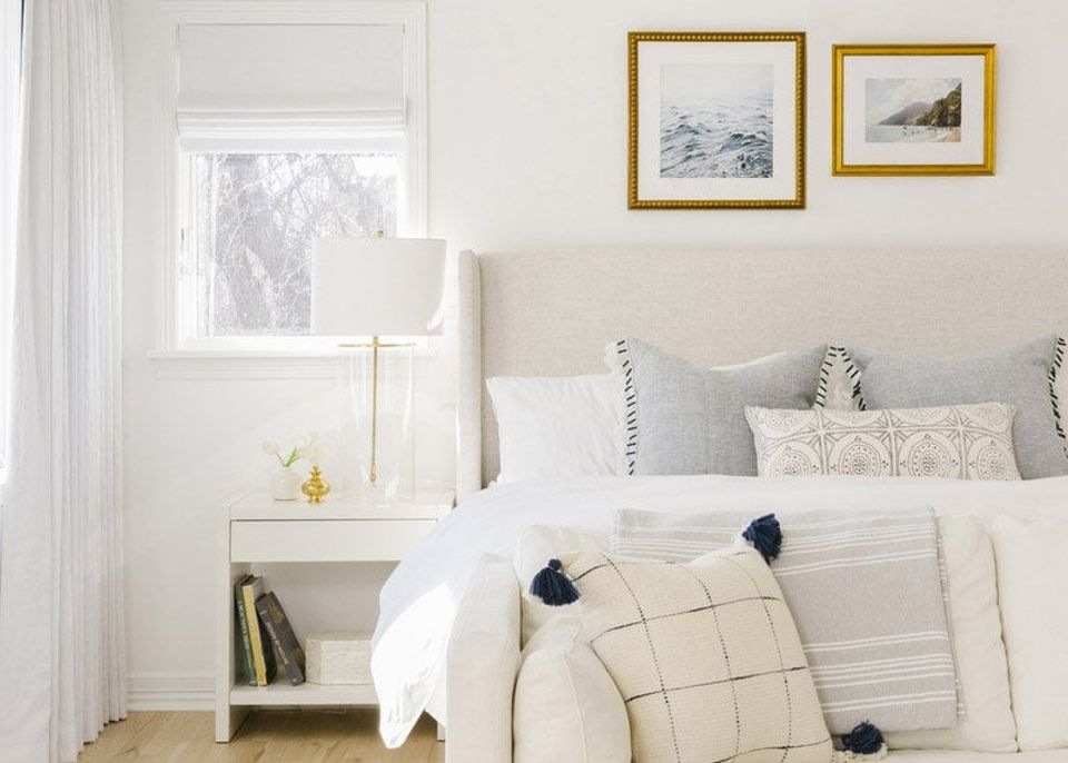 Serene coastal bedroom with cream bedding, white nightstand, brass-framed art, and soft natural light from frosted window