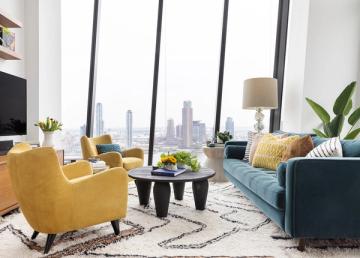 Contemporary urban living room with mustard yellow accent chair, teal sofa, and floor-to-ceiling city views in a sophisticated neutral palette