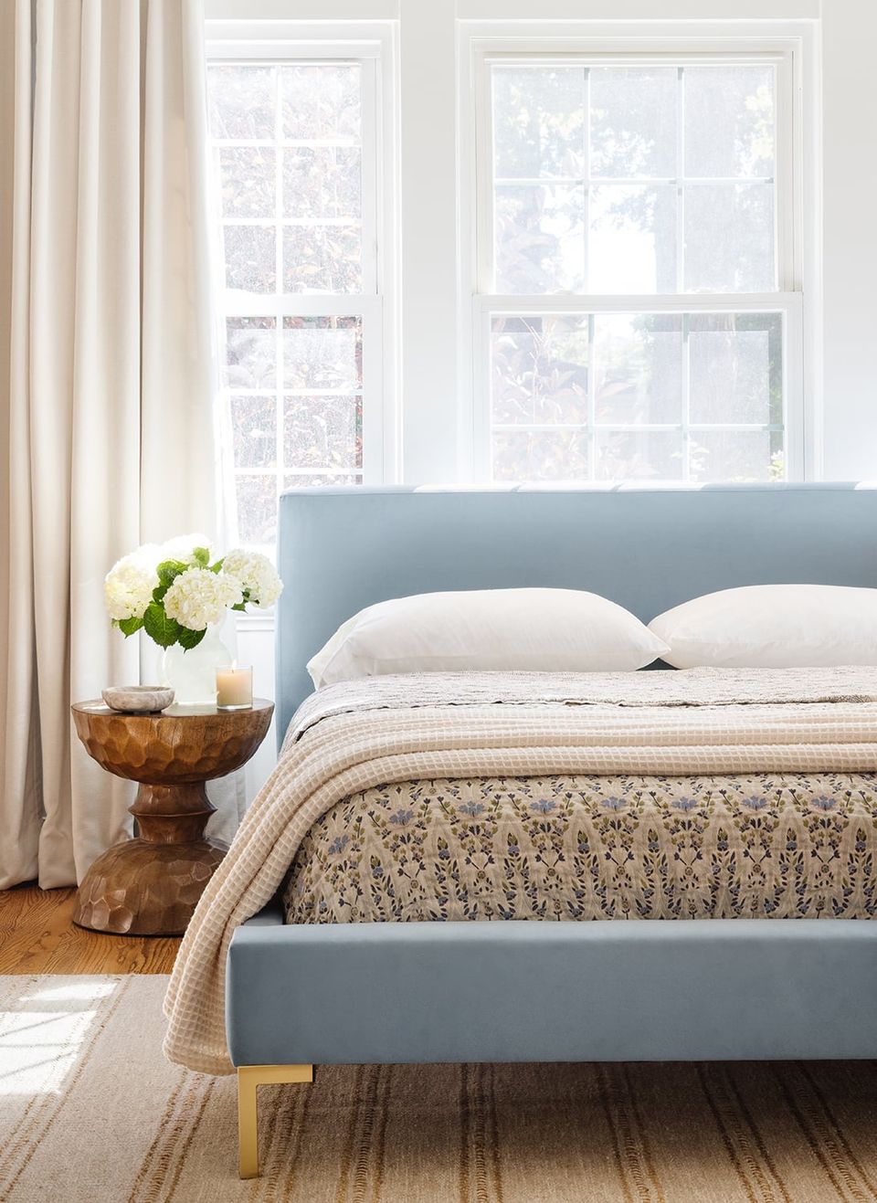 Modern bedroom with soft blue upholstered bed, cream pillows, warm wood accents, and abundant natural light from large windows
