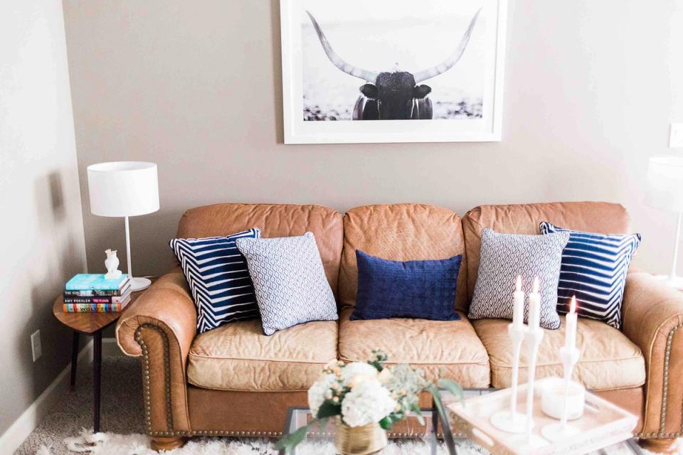 Warm transitional living room with caramel leather sofa, navy and striped pillows, neutral walls, and modern artwork