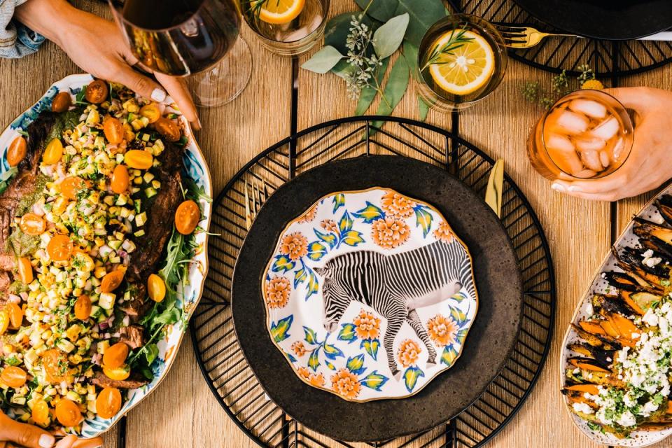 Overhead view of summer BBQ table with vibrant orange and black color palette, featuring patterned dinnerware and fresh seasonal dishes on rustic wood.