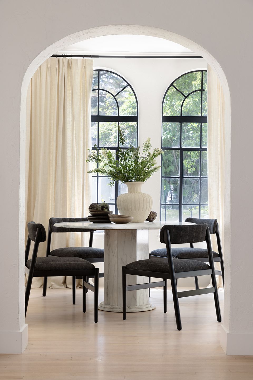Minimalist dining nook with arched entry, black mid-century chairs, cream curtains, and botanical accents creating serene elegance.