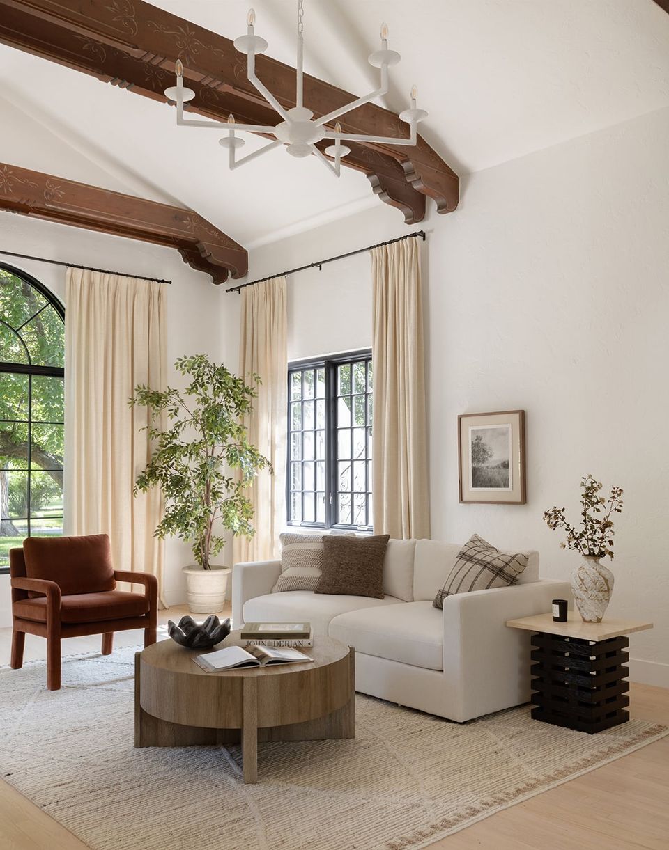 Organic modern living room with exposed wood beams, cream linen sofa, cognac leather chair, and natural wood coffee table against white walls.