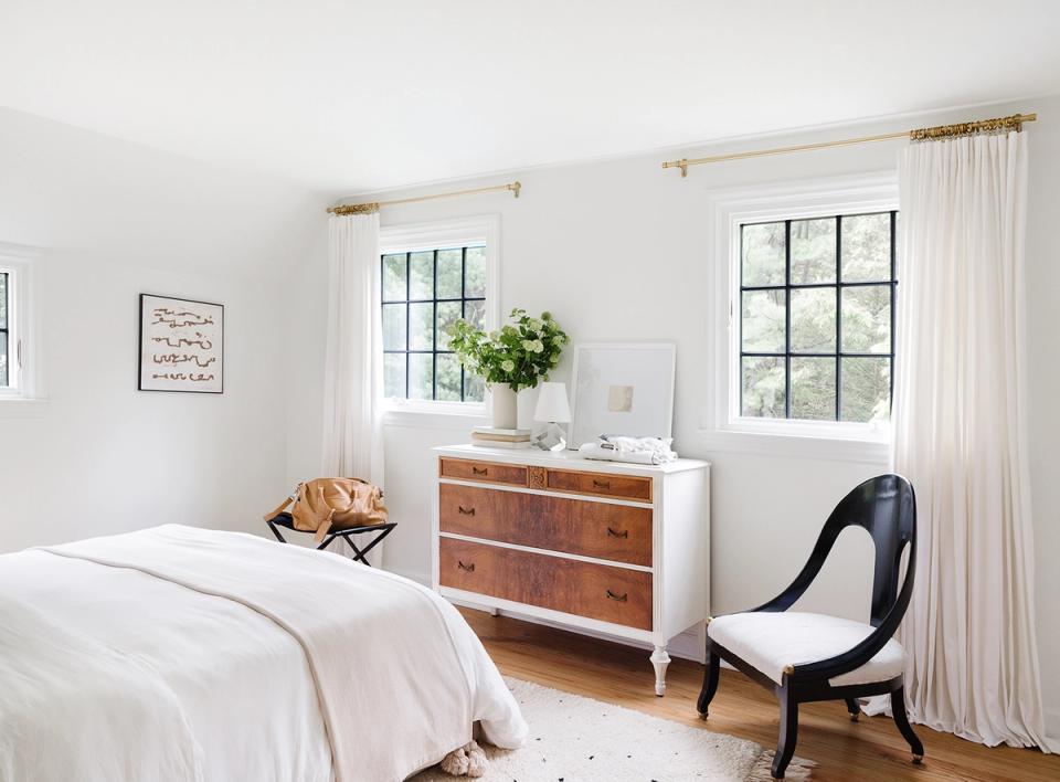 Modern bedroom blending mid-century style with white bedding, warm wood dresser, and sculptural black chair