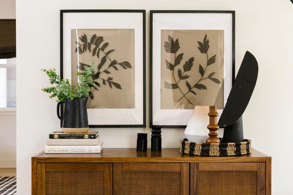 Contemporary eclectic console styling with botanical framed art, warm wood base, black accessories, and fresh greenery in monochromatic backdrop